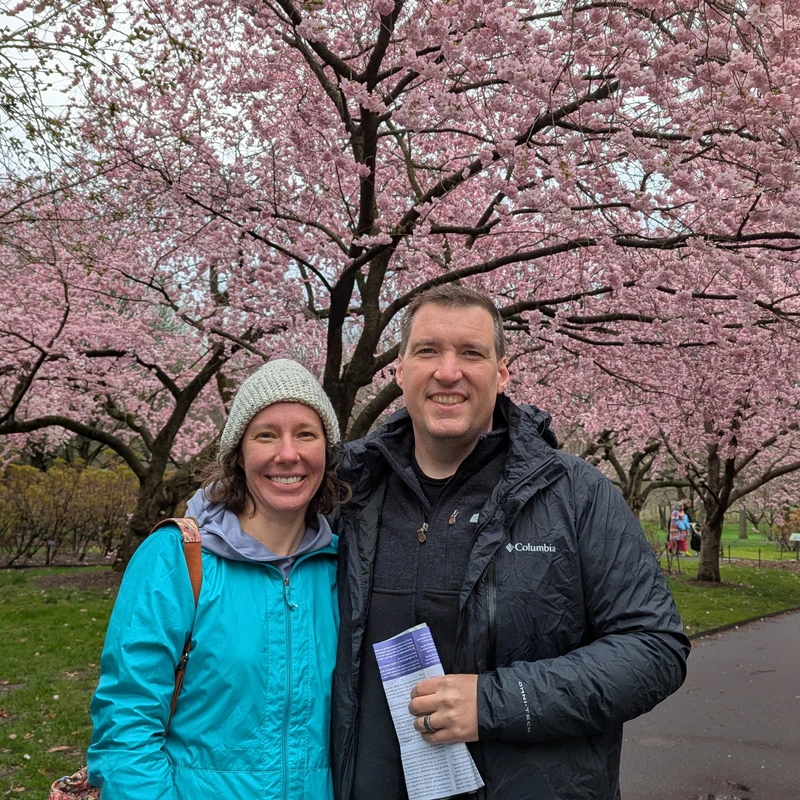A smiling man and woman stand close together in a park. Behind them are large trees covered in beautiful pink cherry blossoms.