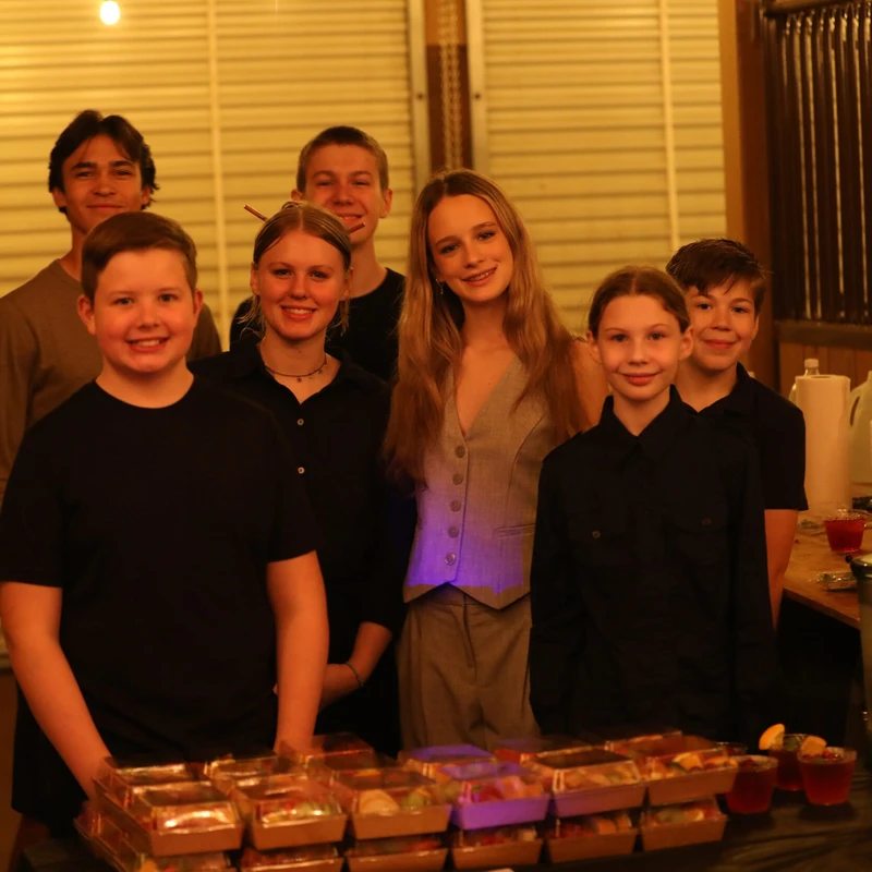 A group of seven smiling teenagers and children stand behind a long table with pre-packaged meals and red drinks at an indoor event with warm lighting.