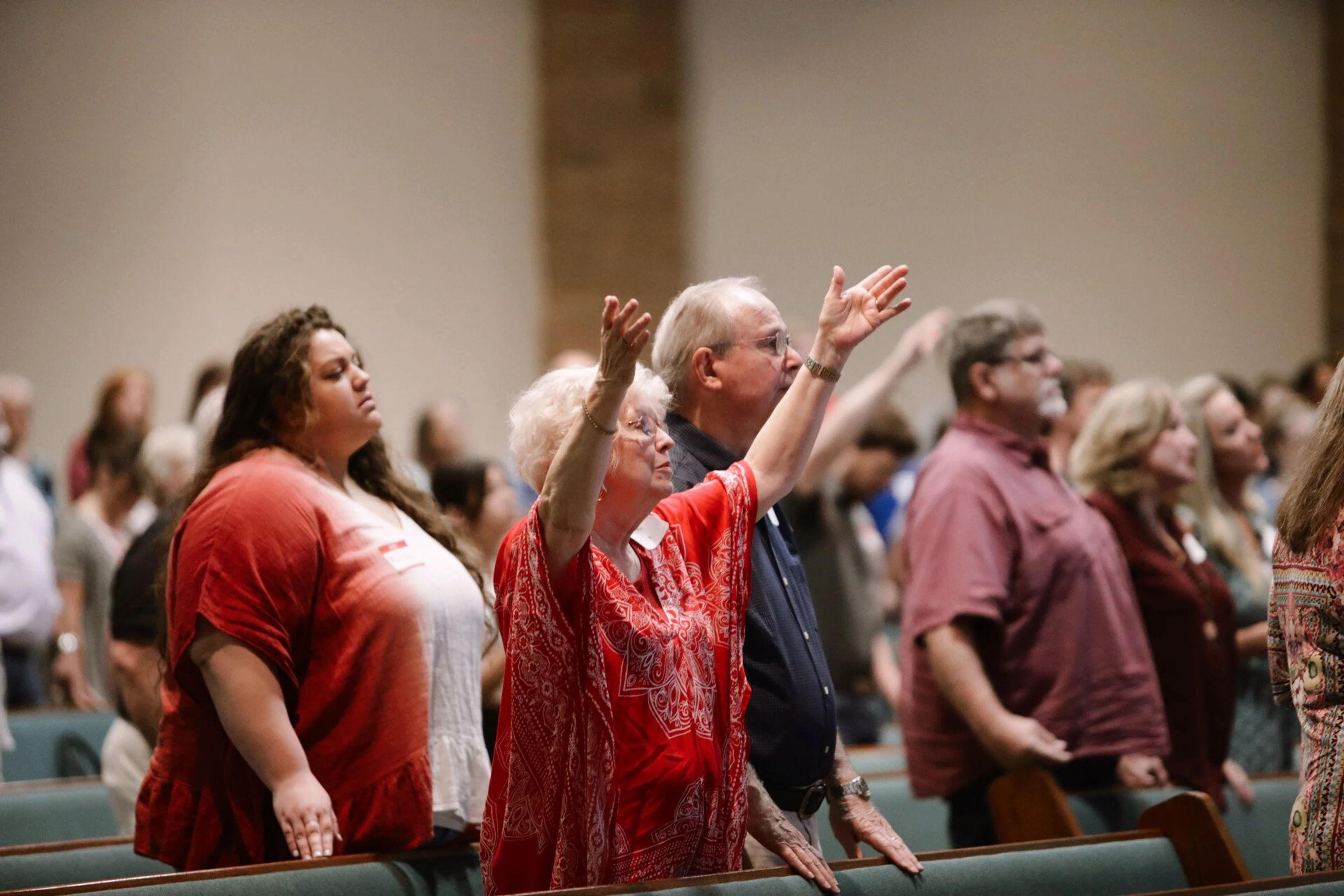 A diverse congregation stands in church pews. An older woman in a red patterned top raises her hands in praise, while others around her also worship.
