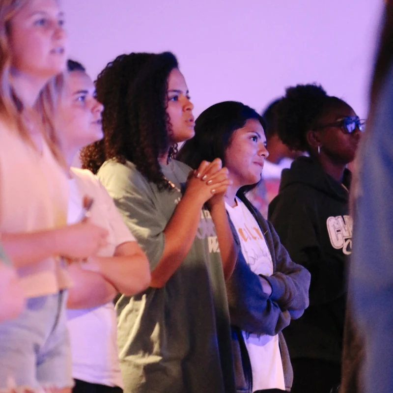 A diverse group of young women stand together in an audience under purple light, looking forward with attentive and engaged expressions.