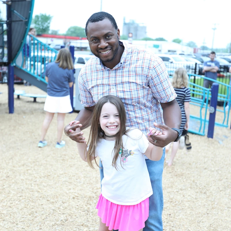 A smiling Black man in a plaid shirt stands behind a young white girl in a pink skirt. He holds her hands as they pose together on wood chips at a playground.