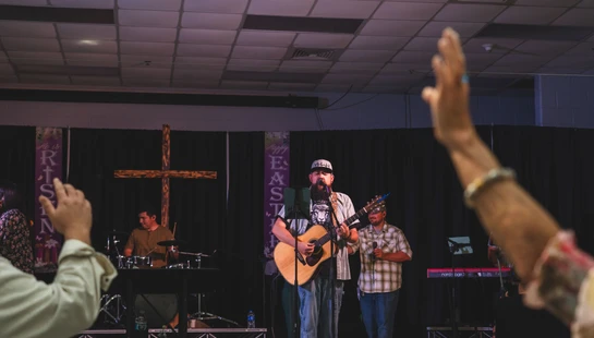 A band with a guitarist and singer leads worship on a stage decorated with a cross and Easter banners, as members of the congregation raise their hands.