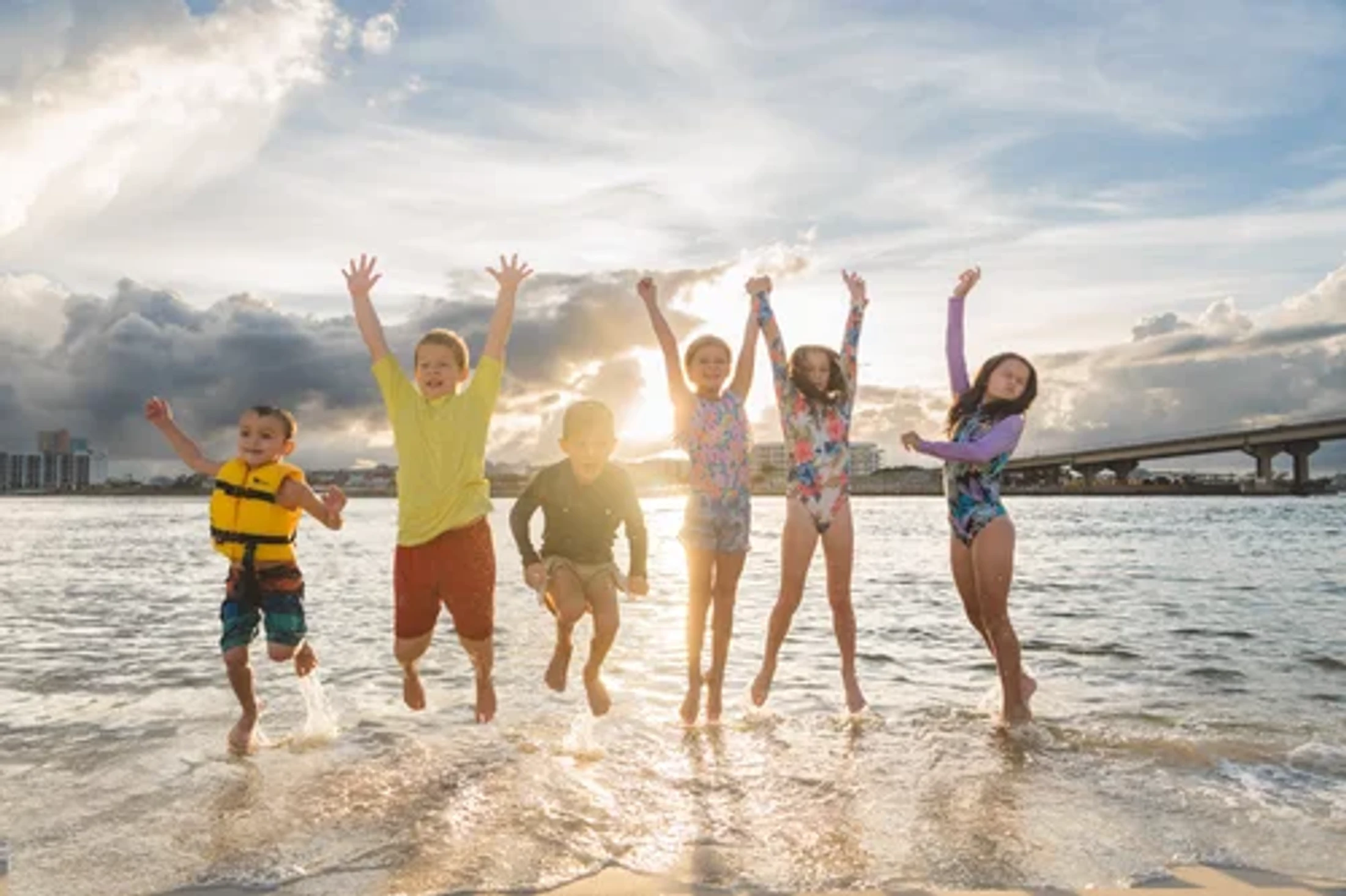 Six happy children in swimwear jump in the shallow ocean water at sunset. A city skyline and bridge are visible in the background.