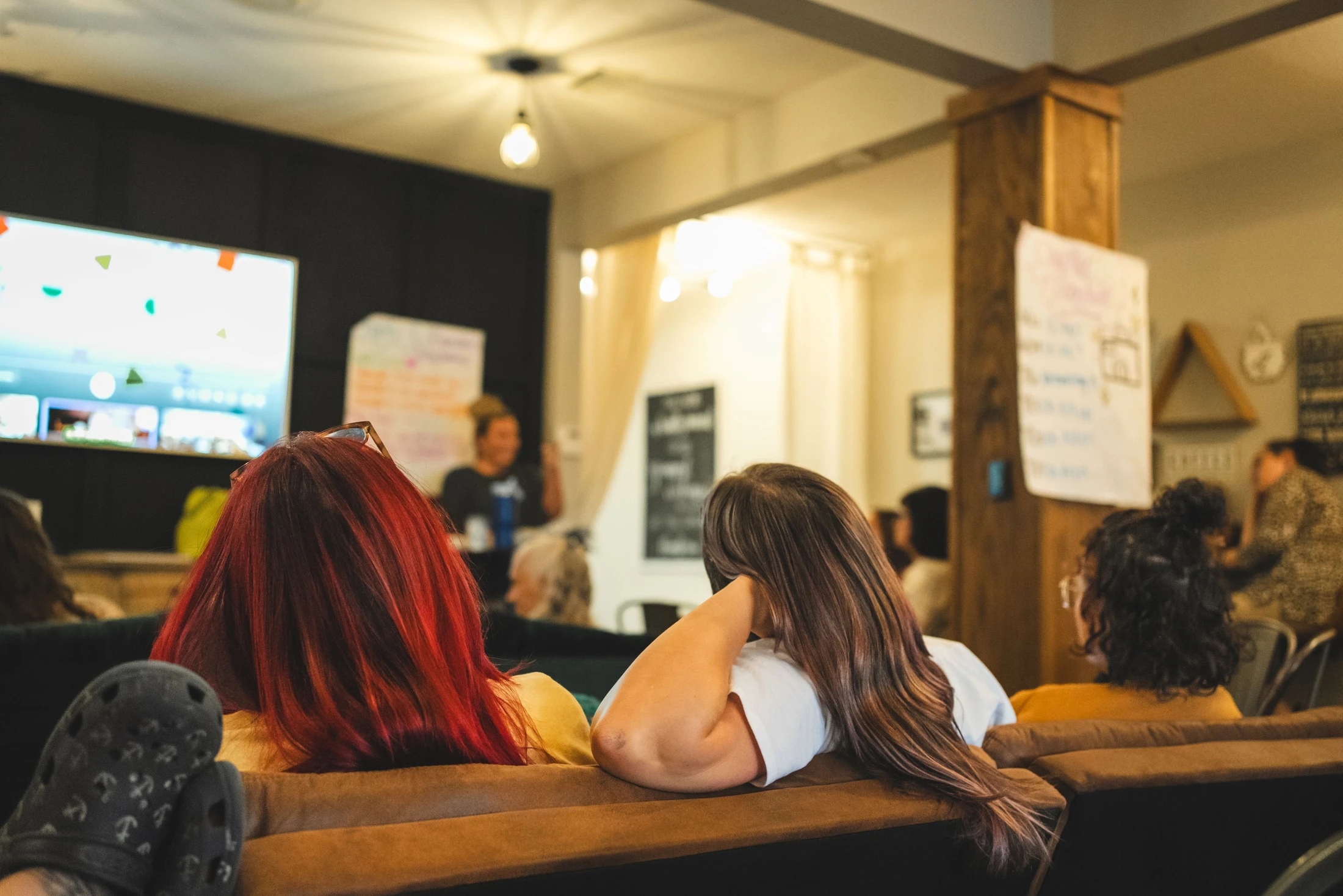 View from behind two women on a couch, watching a speaker give a presentation to a small, seated audience in a cozy, warmly lit room.