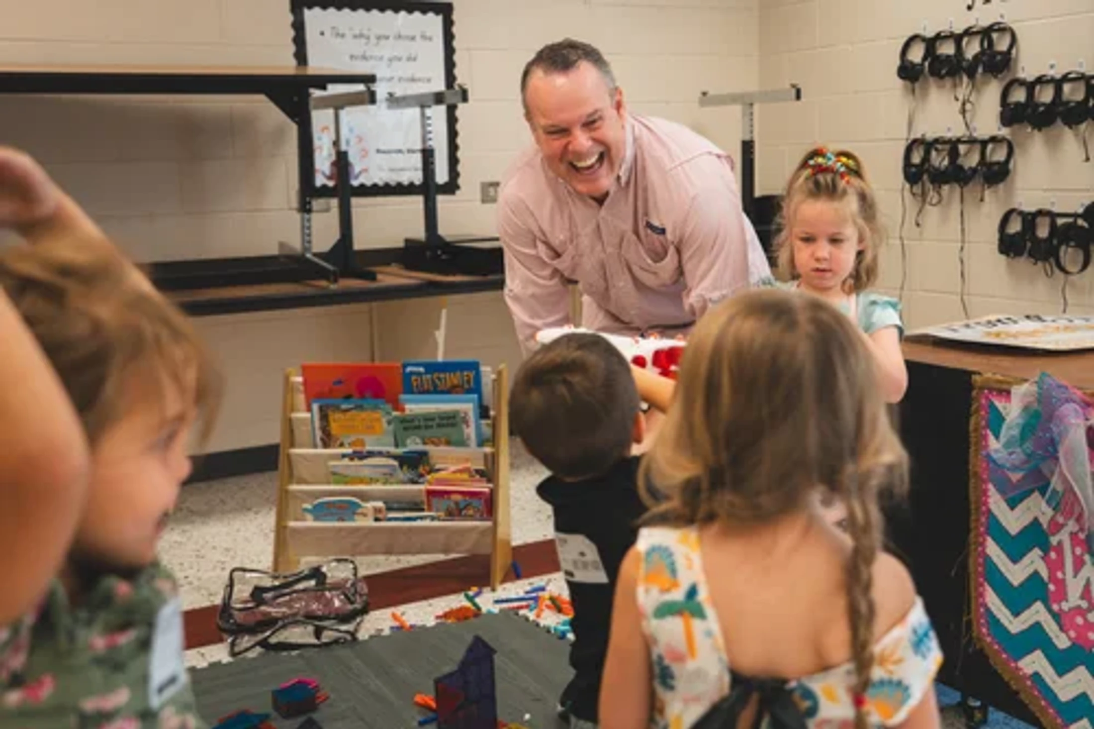 A man in a pink collared shirt laughs heartily as he interacts with a group of young students in their classroom, surrounded by books and learning materials.
