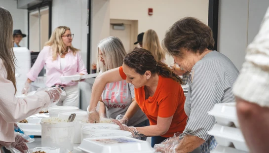 A group of female volunteers stand along a production line, wearing gloves and packing food into takeaway containers for a community outreach event.