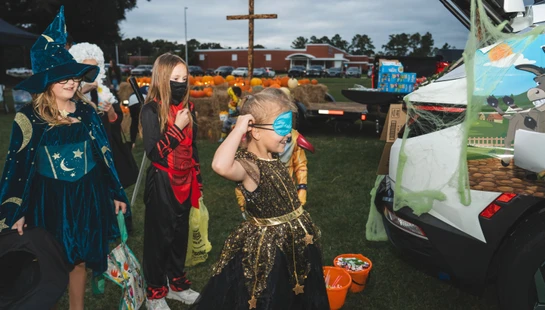 Three children in Halloween costumes gather candy at a trunk-or-treat event held outdoors on a lawn, with a decorated car and pumpkins in the background.
