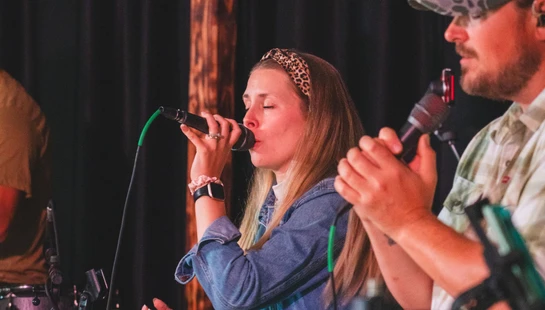 A woman and a man sing passionately into microphones on stage during a performance, with a large wooden cross visible in the background against a black curtain.