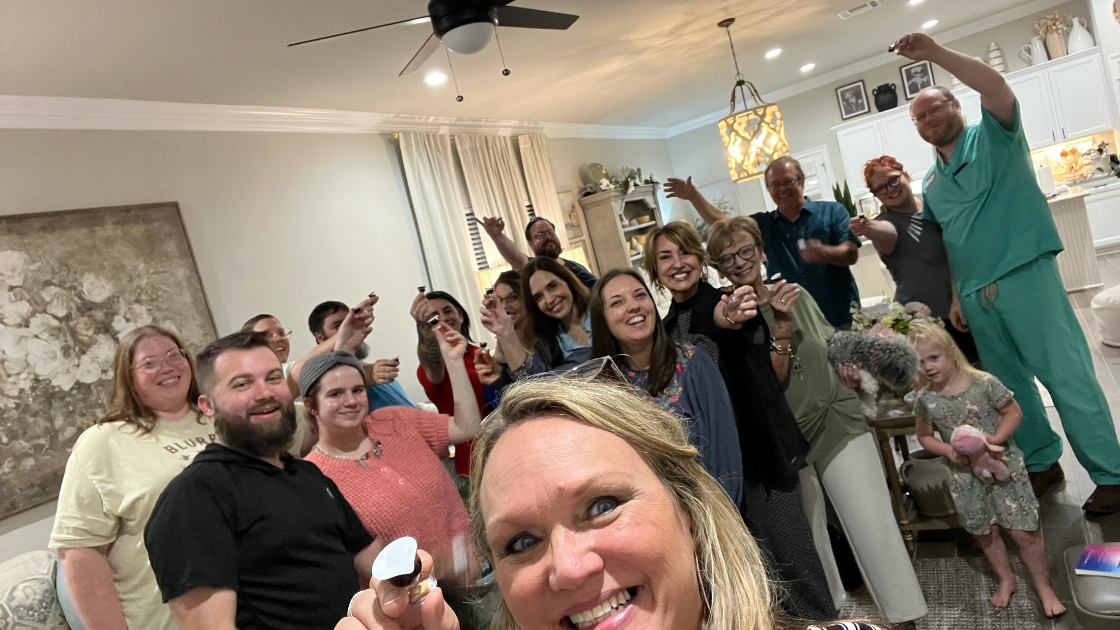 A woman takes a selfie of a large group of smiling friends and family gathered in a living room, many holding up small communion cups in celebration.