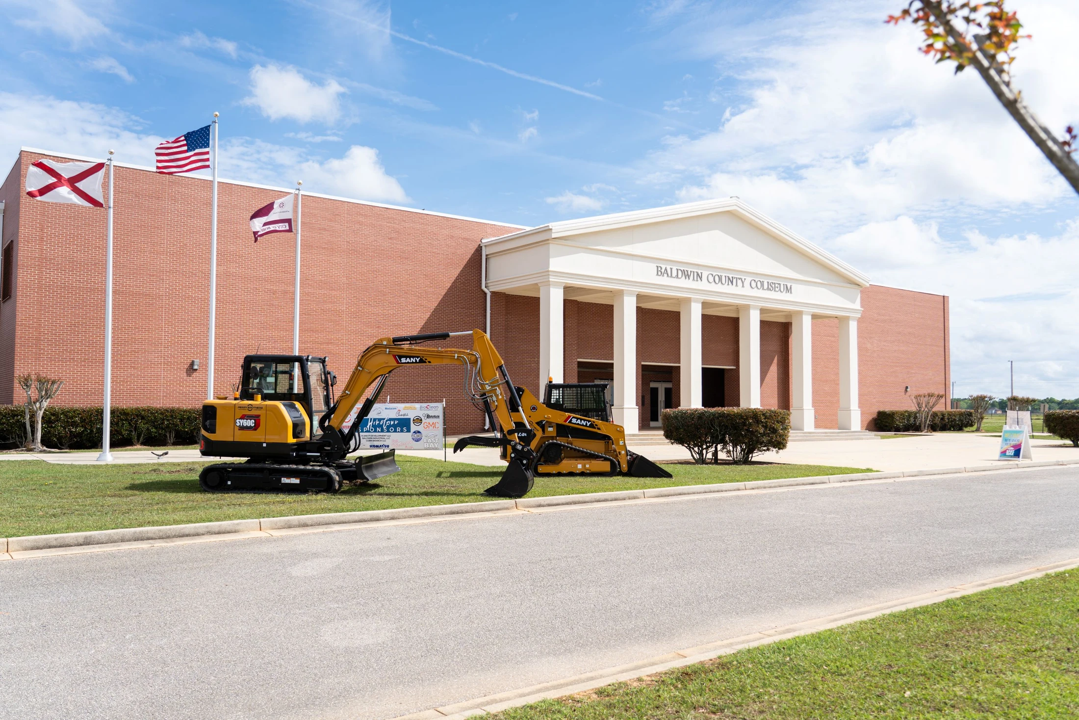 A yellow excavator and skid steer loader are parked on the lawn in front of the red brick Baldwin County Coliseum under a bright blue sky with clouds.