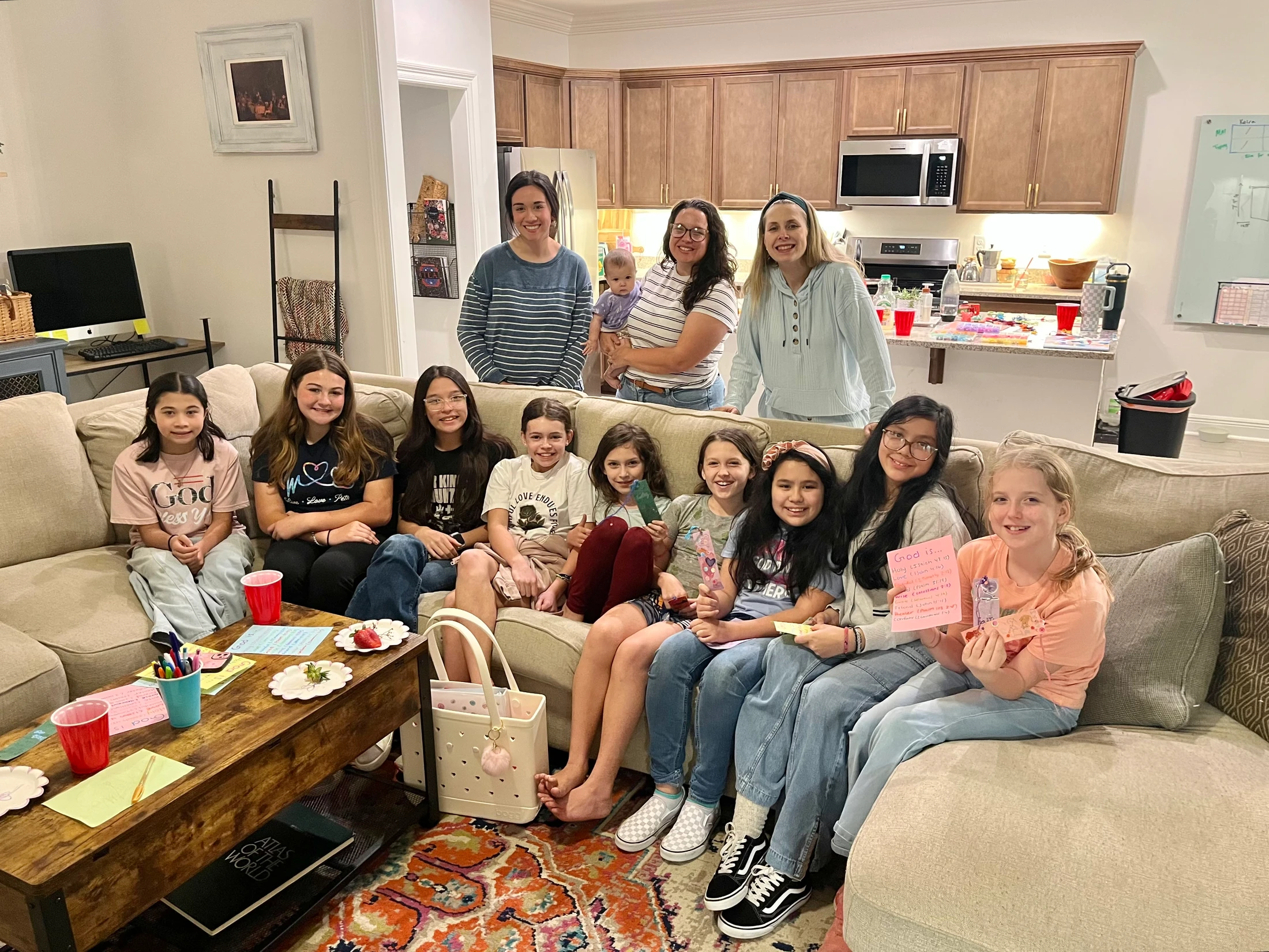 A large group of smiling girls and women pose for a photo on a couch in a living room, with craft supplies on the coffee table in front of them.