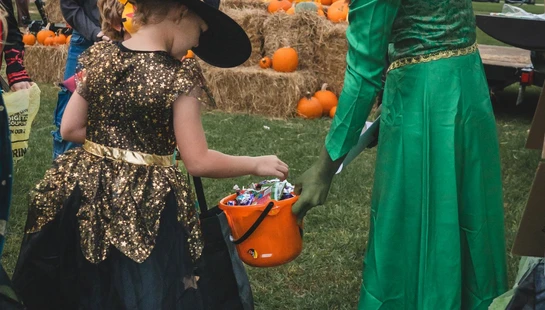 A young girl in a witch costume receives candy in her orange bucket from a person dressed as Princess Fiona at an outdoor fall festival with pumpkins.