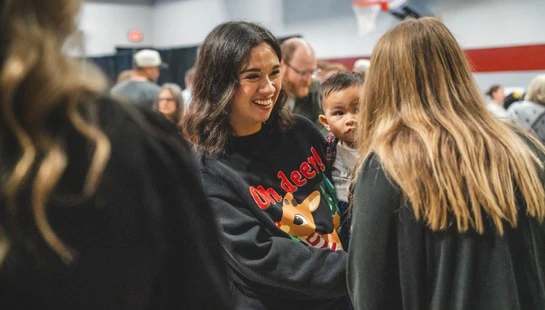A smiling woman in a black 'Oh deer!' sweater holds a small child while talking with another person in a crowded room. A basketball hoop is in the background.