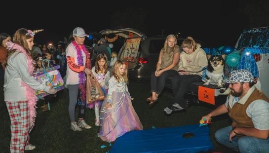 A group of adults and children in various costumes gather at a nighttime trunk or treat event, playing games and collecting candy next to decorated cars.