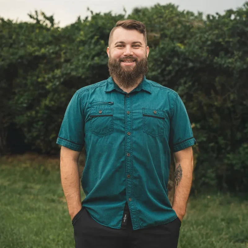 A man with a brown beard and a teal button-up shirt stands in front of a hedge, smiling at the camera with his hands in the pockets of his black shorts.