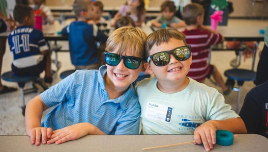Two young boys sit at a desk, smiling for the camera. They both wear novelty sunglasses with 'VBS 2026' written on the lenses in a busy classroom.