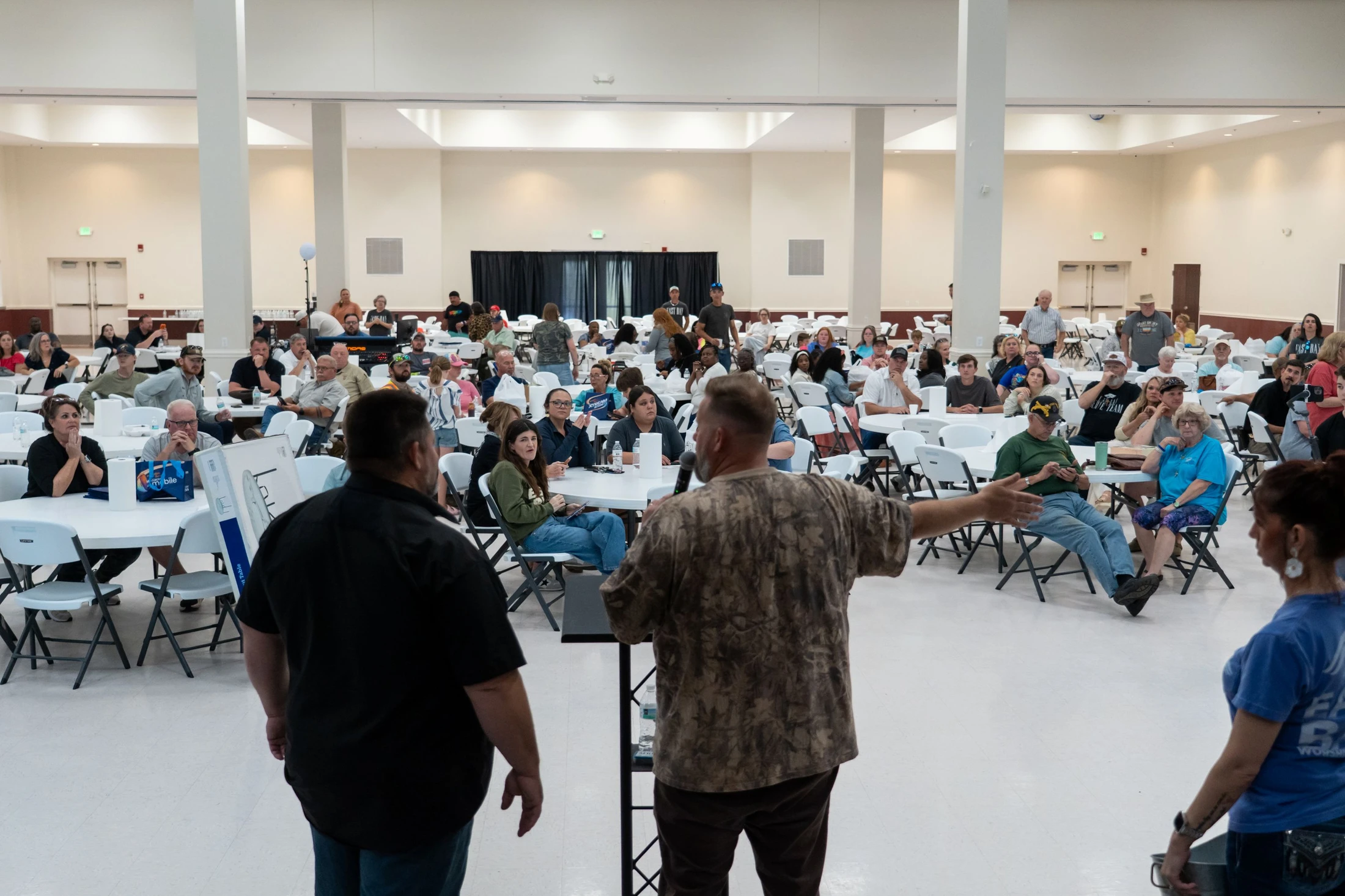 A man in a camo shirt addresses a large crowd seated at tables in a brightly lit community hall during an event.