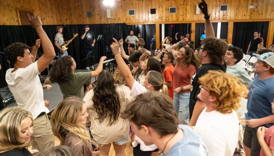 A large group of young people dance and sing with their hands in the air during a live concert in a rustic wooden hall.