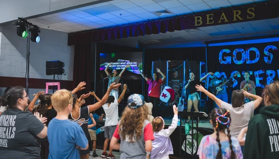 A crowd of young people with their hands raised, watching a band lead a worship service on a blue-lit stage with lyrics displayed on a screen.