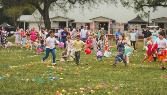 A large group of excited children run across a grassy field to collect colorful plastic eggs during an Easter egg hunt, as adults watch in the background.