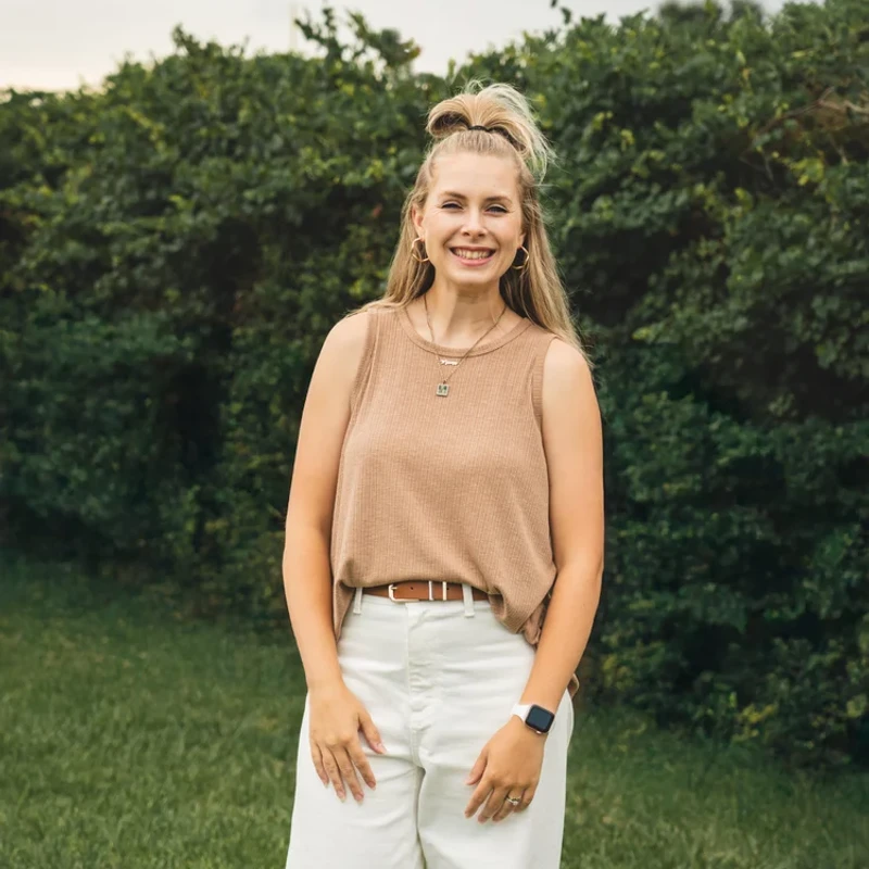 A smiling blonde woman with a half-up bun hairstyle poses in a grassy field. She wears a brown sleeveless top, white pants, and gold jewelry.