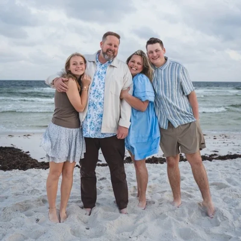 A family of four, a mother, father, son, and daughter, stand close together and smile for a photo on a sandy beach with the ocean behind them.