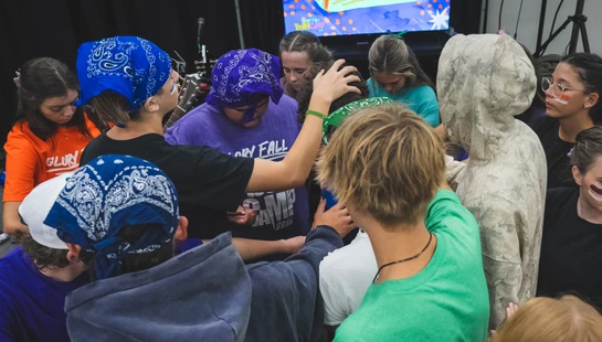 A group of young people at a youth camp gathered in a supportive circle, placing their hands on a person in the center in a moment of prayer.