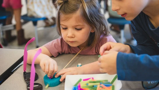 A young girl and a boy with a hearing aid sit closely at a table, focused on an arts and crafts project involving colorful sticks and blue modeling clay.