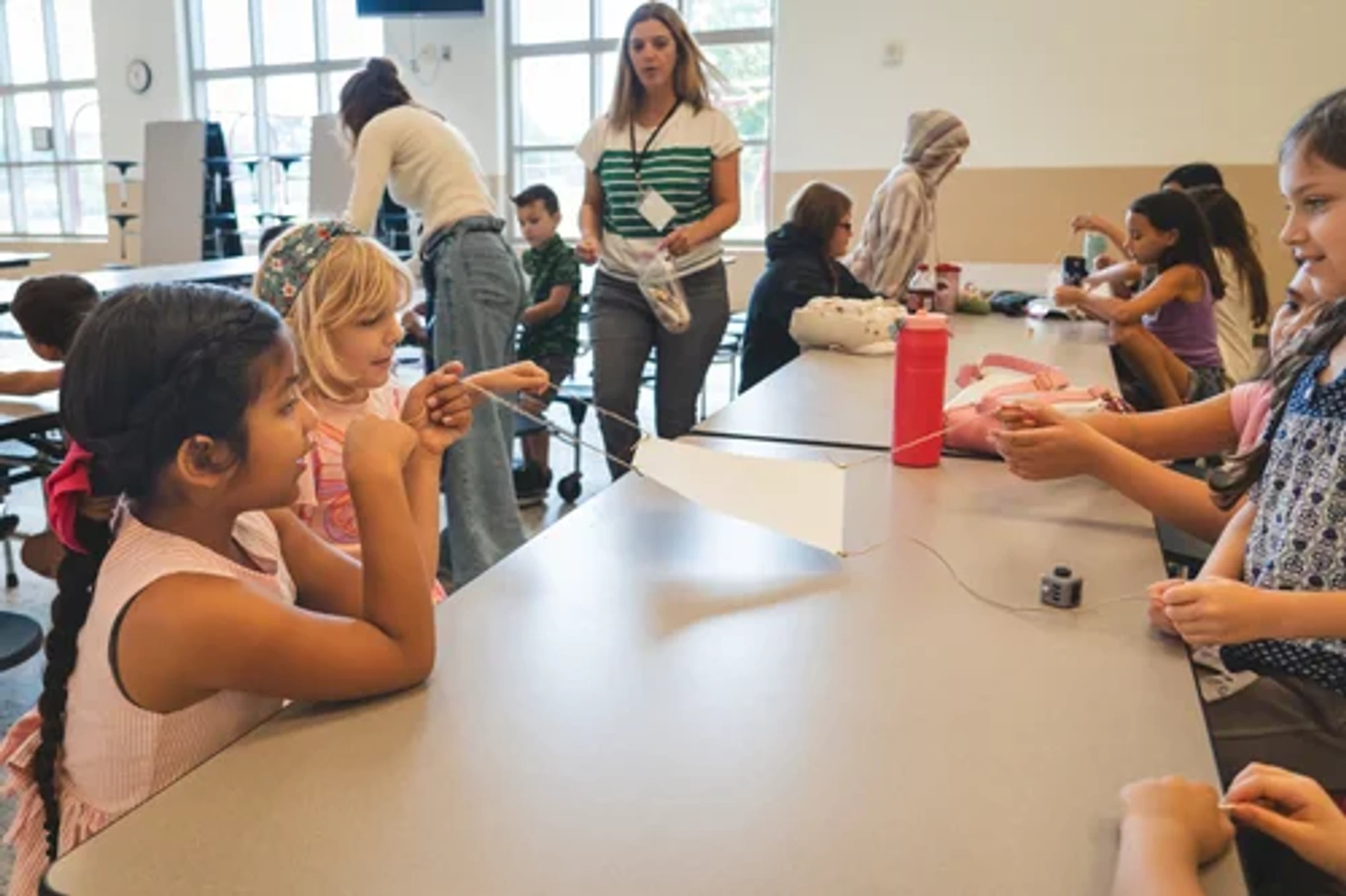 A group of elementary school students sits at a long table in a classroom, engaged in a hands-on learning project with strings as a teacher supervises.