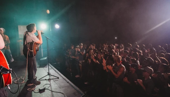 A musician with an acoustic guitar sings into a microphone on stage, illuminated by spotlights, as a large audience claps and watches from the dark.