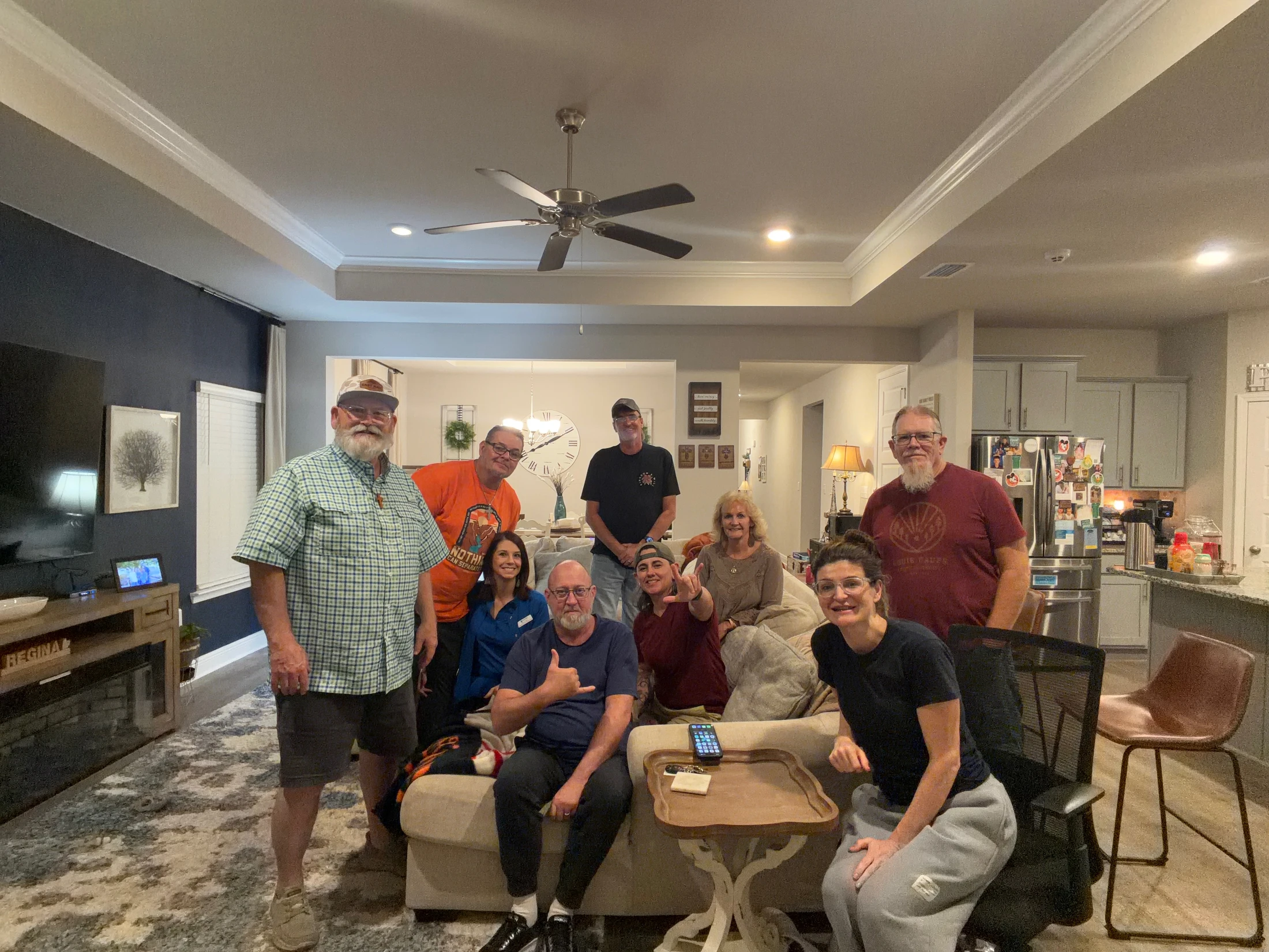 A group of nine adults of various ages smile and pose for a photo together in a modern, well-lit living room, gathered around a light-colored couch.