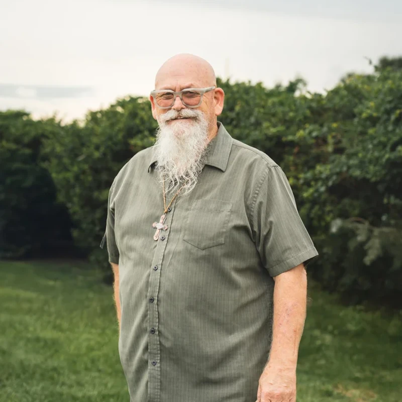 A bald, older man with a long white beard and glasses smiles warmly. He is standing in a grassy area wearing a green shirt and a prominent cross necklace.