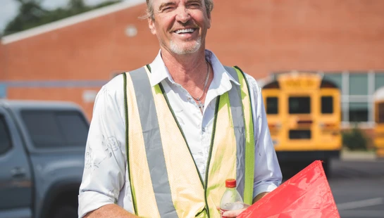 A smiling man wearing a yellow safety vest stands in a school parking lot, holding a red flag and a bottle of soda, with a school bus in the background.