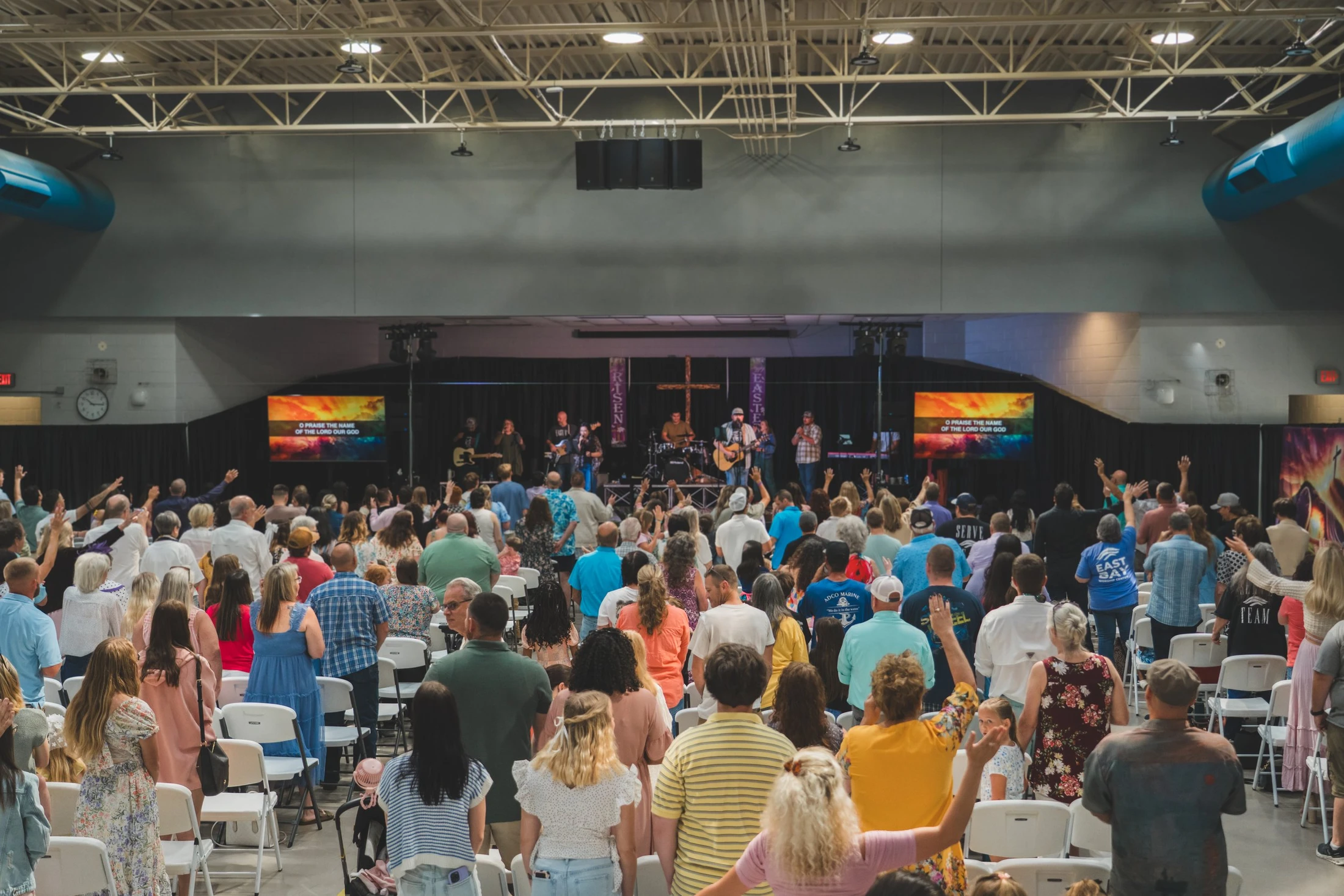 A large congregation seen from behind, standing and raising their hands in worship towards a band on a stage during a contemporary church service.