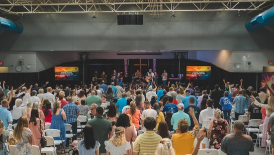 A large congregation seen from behind, standing and raising their hands in worship towards a band on a stage during a contemporary church service.