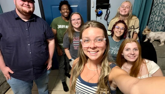 A woman takes a selfie with seven smiling friends gathered in an entryway. A small white dog is visible on a couch to the right.