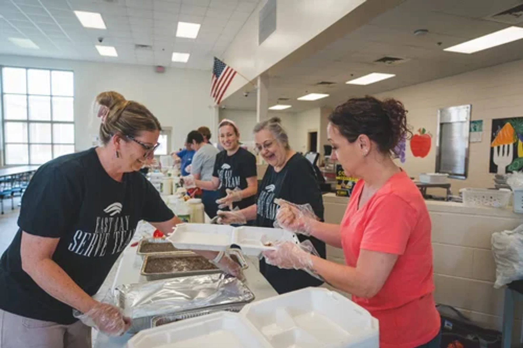 A group of smiling female volunteers wearing gloves serves food from large trays into takeout containers in a brightly lit cafeteria.