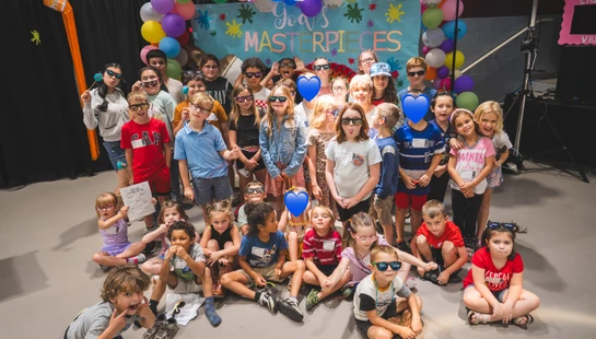 A large group of children pose for a fun photo with silly faces and sunglasses under a colorful balloon arch and a banner reading 'God's Masterpieces'.