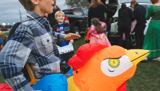 A young boy in a plaid shirt wears a funny inflatable rooster costume while trick-or-treating outdoors at a community event with other families.