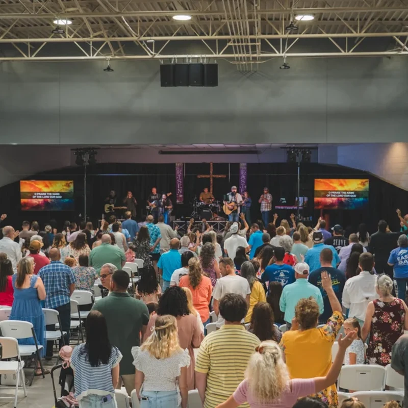 A large congregation stands in a hall, facing a stage where a band is leading worship. Many people have their hands raised in praise.