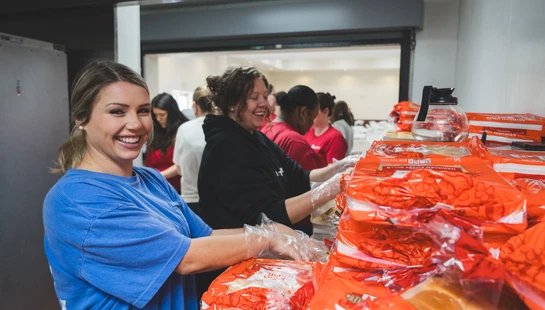 A smiling woman in a blue shirt looks at the camera while preparing food with other volunteers in a kitchen. Stacks of bread rolls are on the counter.