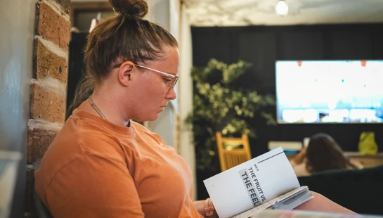 A woman with brown hair in a bun and wearing glasses sits in profile reading a book. She wears an orange t-shirt and is focused on the page.