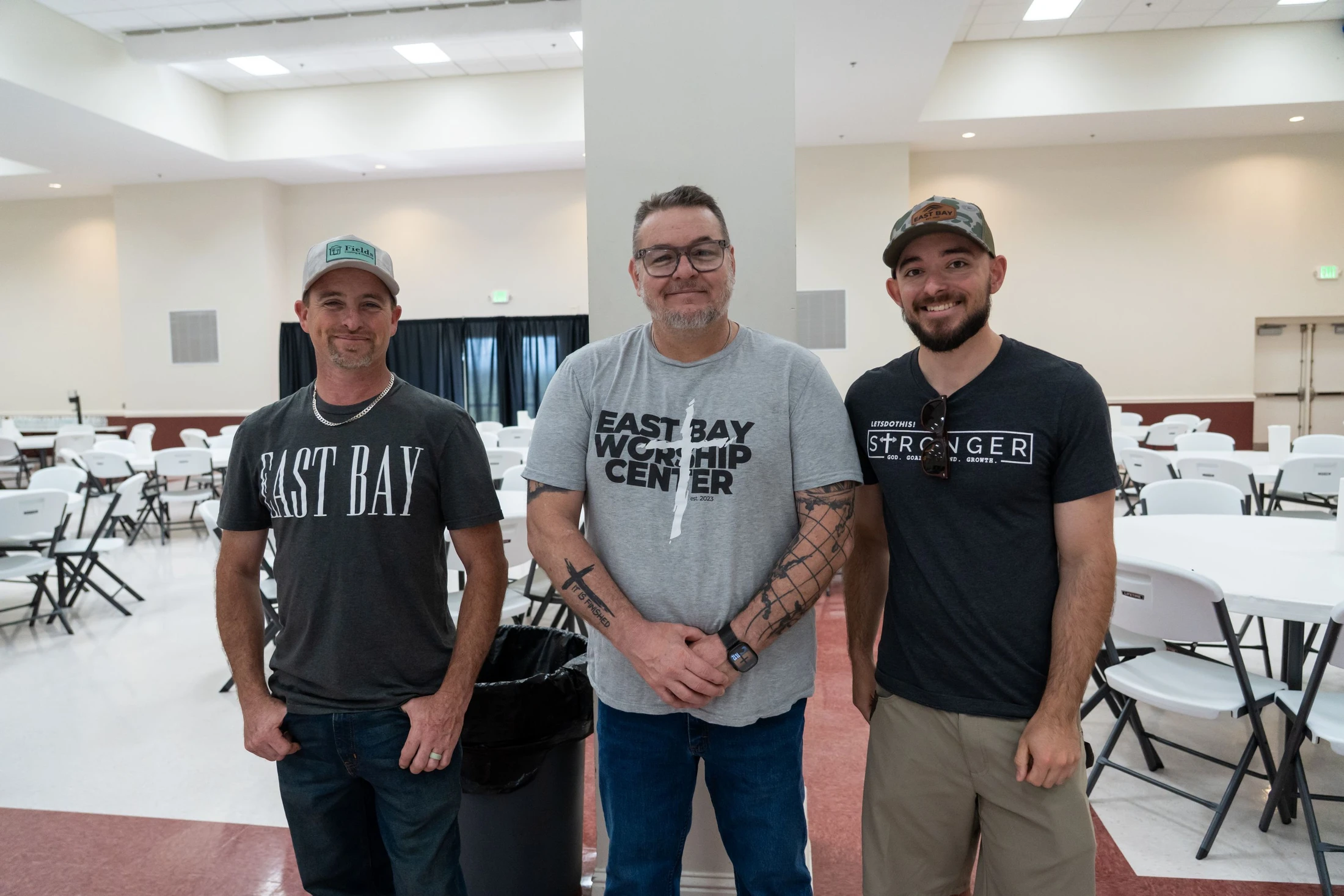 Three men are standing together and smiling for a photo inside a large hall with many empty white tables and chairs in the background.