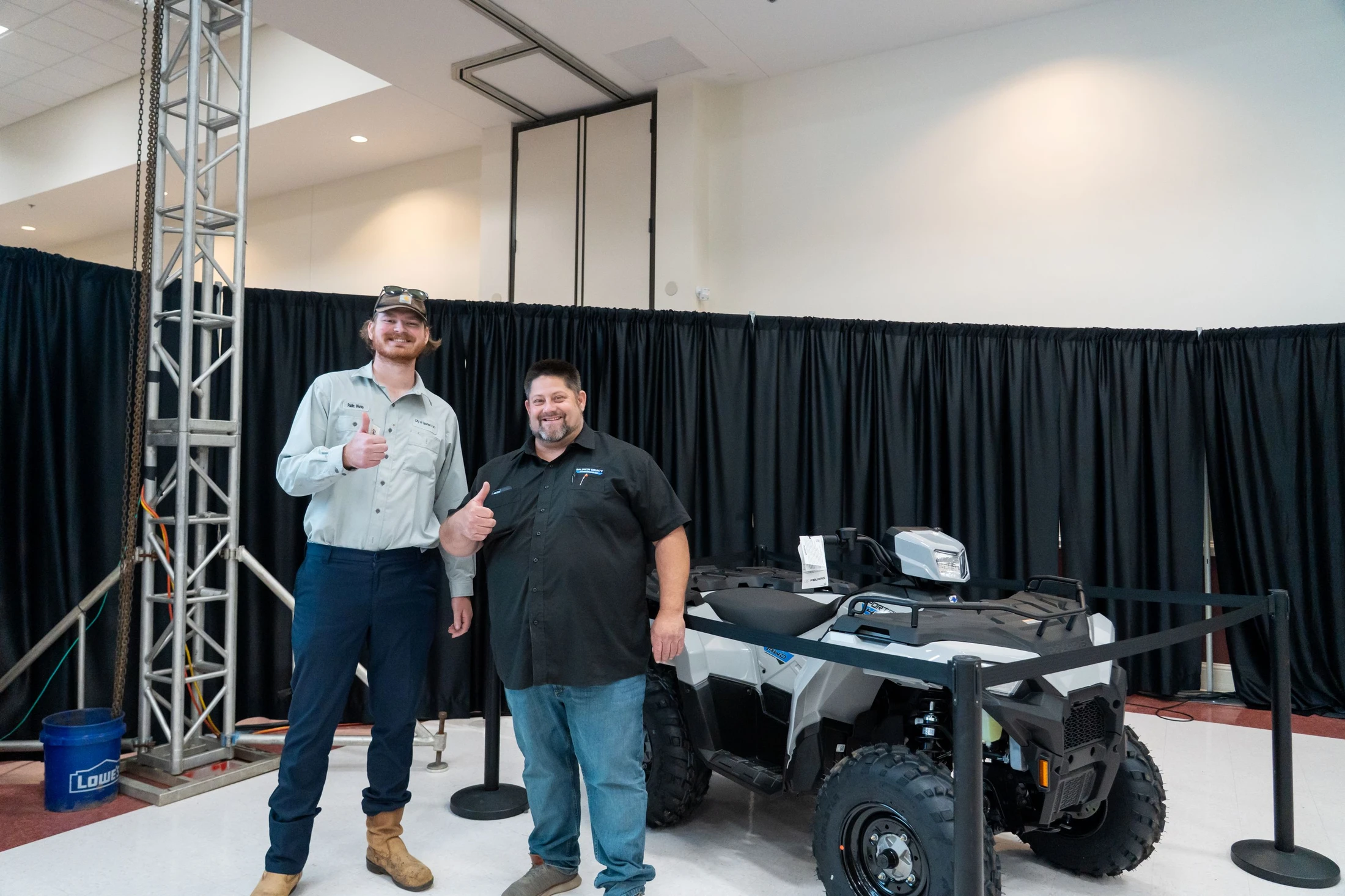 Two men smile and give a thumbs-up while standing next to a new white Polaris all-terrain vehicle on display indoors in front of a black curtain.