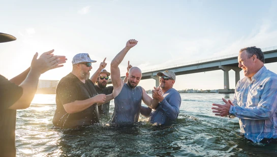 A group of men celebrate as a man raises his fist triumphantly during a water baptism ceremony, with a sunny sky and bridge in the background.