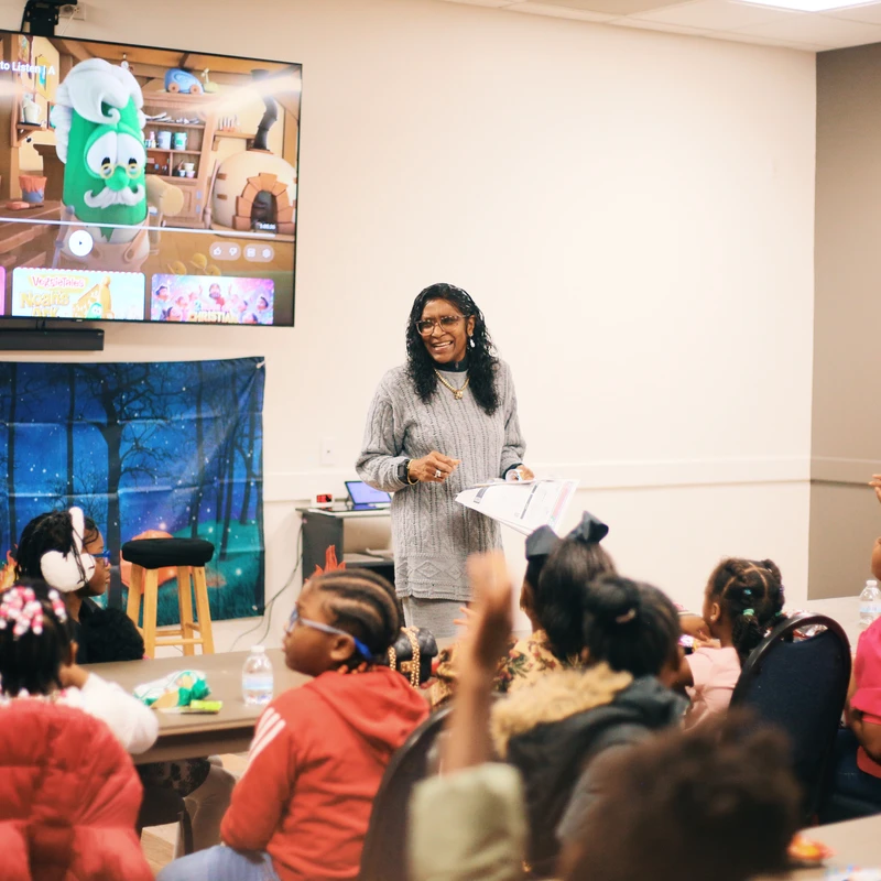 A smiling woman stands at the front of a room, teaching a group of young children sitting at tables. Behind her, a VeggieTales cartoon plays on a TV screen.