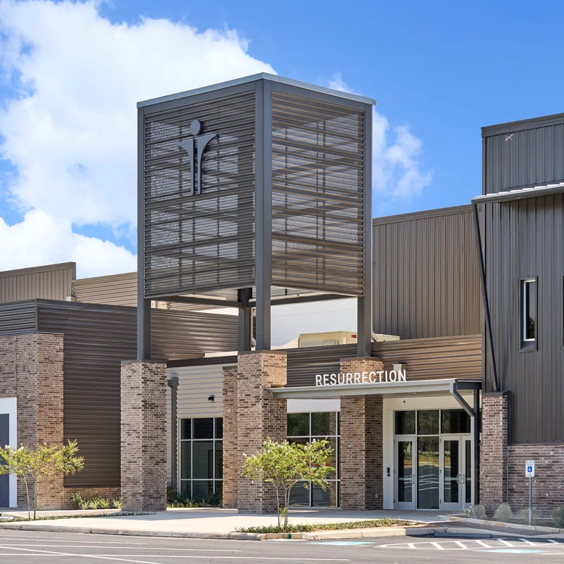 The exterior of Resurrection Church, a modern building with brown brick, dark metal siding, and a central tower with a logo, under a blue sky.