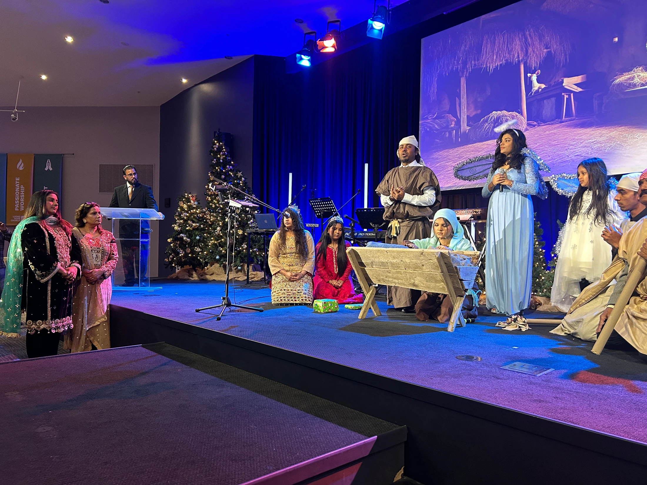 A diverse group of adults and children perform a nativity play on a stage with blue lighting. They are in costume as Mary, Joseph, and angels around a manger.