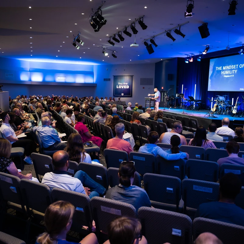 A wide view of a large congregation seated in an auditorium, listening to a pastor on a blue-lit stage during a church service.