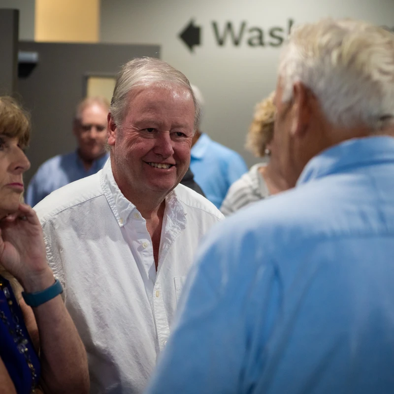 A man in a white shirt smiles while talking to another man whose back is to the camera. A woman in blue stands beside him, listening intently.